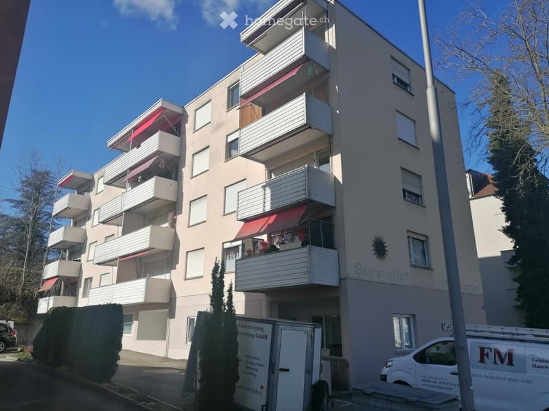 Multi-story residential building with balconies, beige facade, and red awnings. The building is named "Sunnehof" and has a sign on the side.