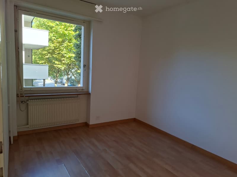Empty room with wooden flooring, white walls, and a large window with a view of trees and a building outside. A radiator is visible below the window.