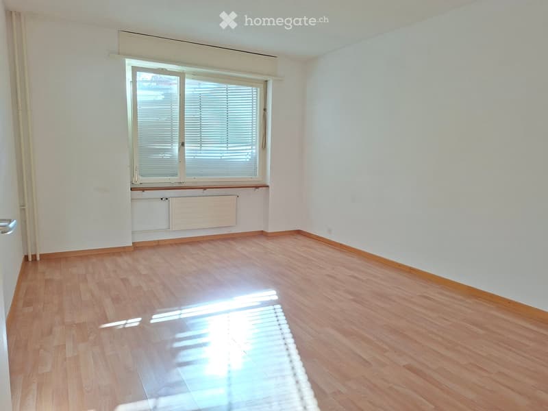 Empty room with wooden flooring, white walls, and a large window with blinds. The room has a radiator below the window and a door visible on the left side.