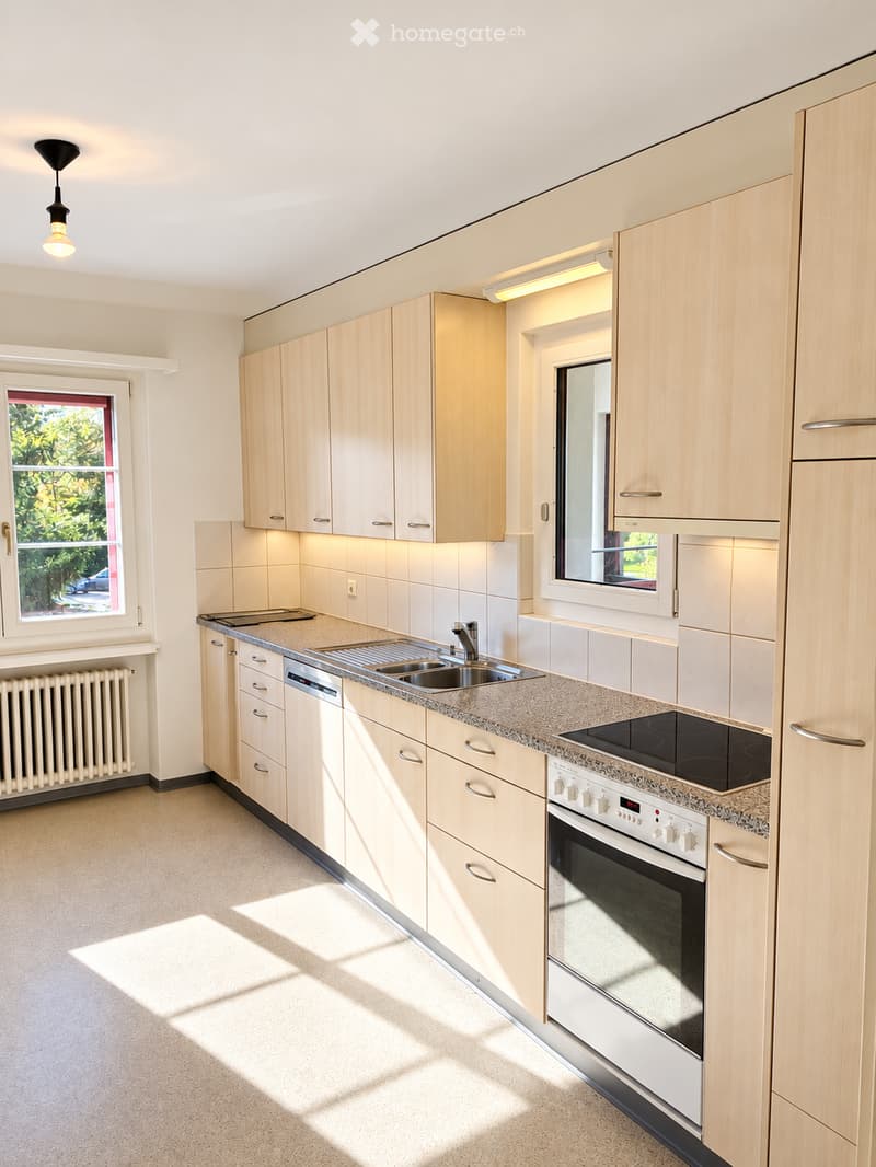 Modern kitchen with light wooden cabinets, granite countertop, stainless steel sink, oven, and stove. Features large windows with a view of greenery outside.