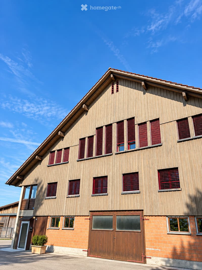 A large wooden barn with red shutters and a pitched roof, featuring a large wooden double door and several windows.