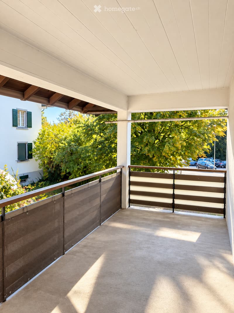 Balcony with a concrete floor, white wooden ceiling, and metal railings with brown and white panels. The view includes green trees and a street with parked cars.