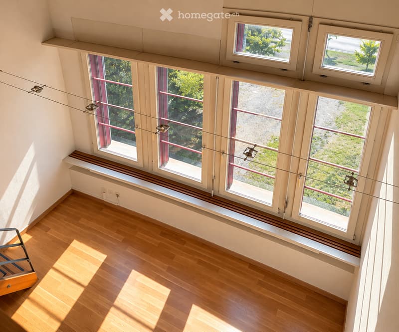 Bright, empty room with wooden flooring, large windows, and modern lighting fixtures suspended from the ceiling.