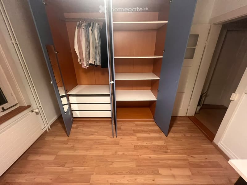 Empty room with wooden flooring, open closet with shelves and drawers, and a window on the left side.