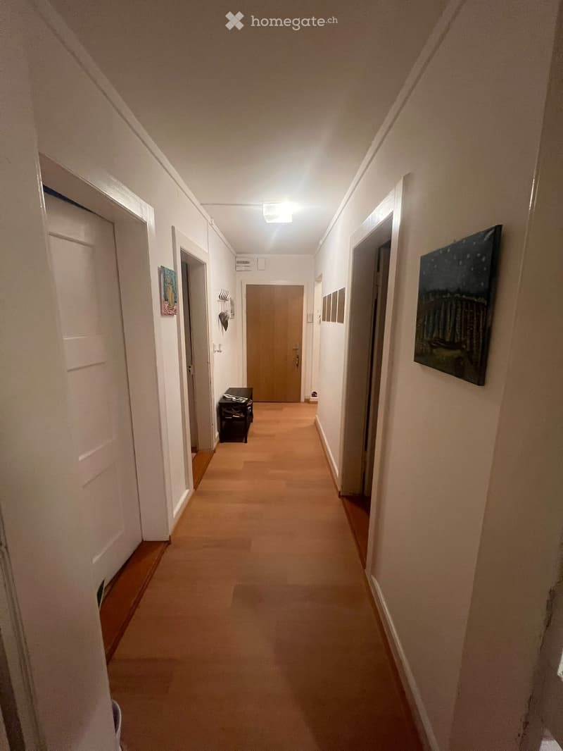 A narrow hallway with light-colored walls, wooden flooring, and several doors leading to different rooms. There are small black tables and artwork on the walls.