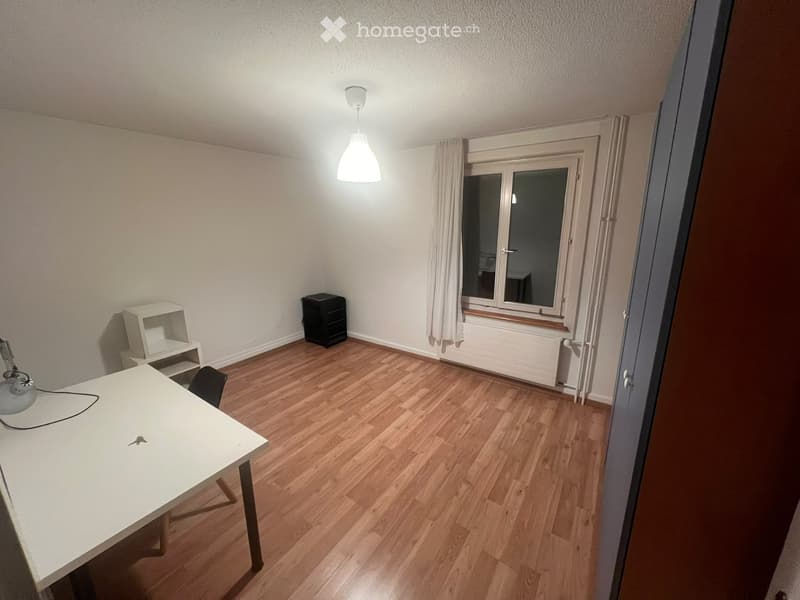 A small, unfurnished room with wooden flooring, a white table, a chair, a small shelf, and a lamp.