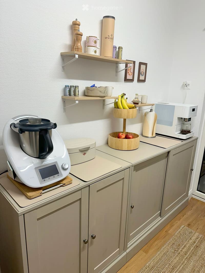 The kitchen features light-colored cabinets, a wooden countertop, and various appliances including a coffee maker, toaster, and blender. There are shelves with cups, bowls, and fruits, as well as framed pictures on the wall.