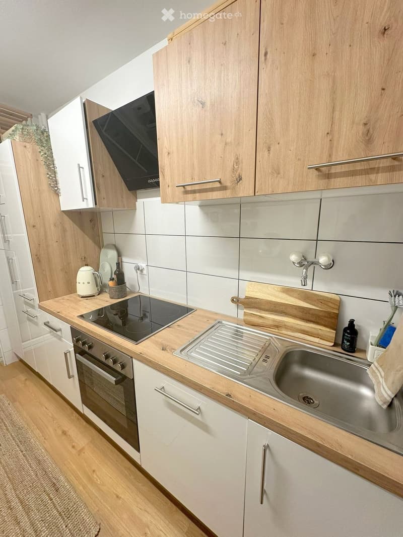 Modern kitchen with light wooden cabinets, white tiles, stainless steel sink, induction stove, and various small appliances.