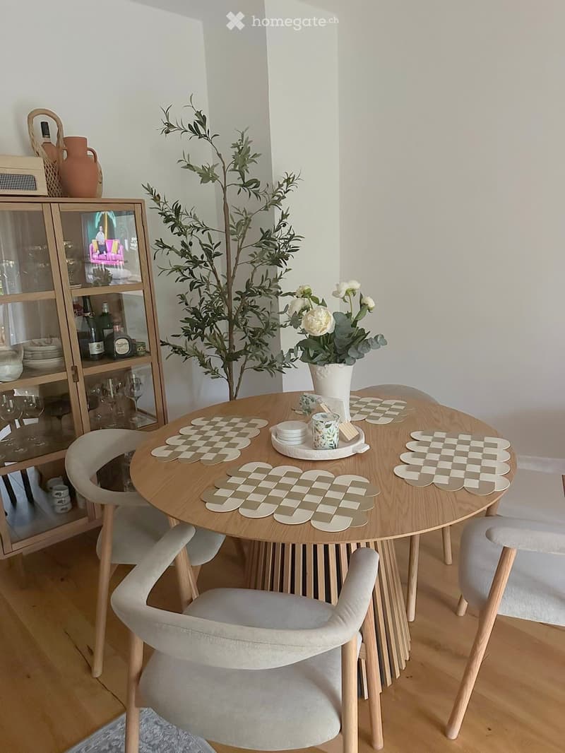 A modern dining area with a round wooden table, four light grey upholstered chairs, a flower vase with white roses, and a wooden cupboard with glass doors displaying wine glasses and bottles.