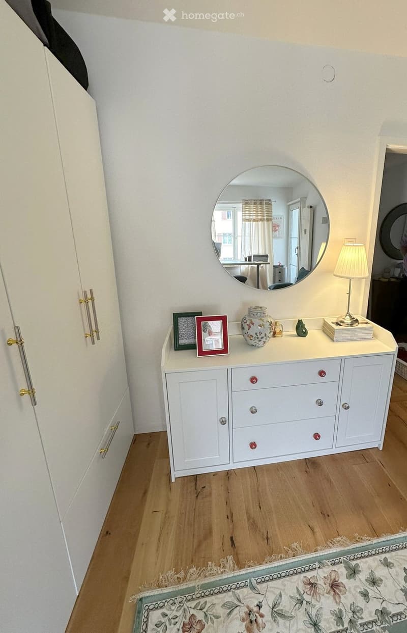 A well-lit hallway with a white dresser, a round mirror above it, a table lamp, picture frames, a decorative vase, and books on the dresser. Wooden flooring and a patterned rug are visible.