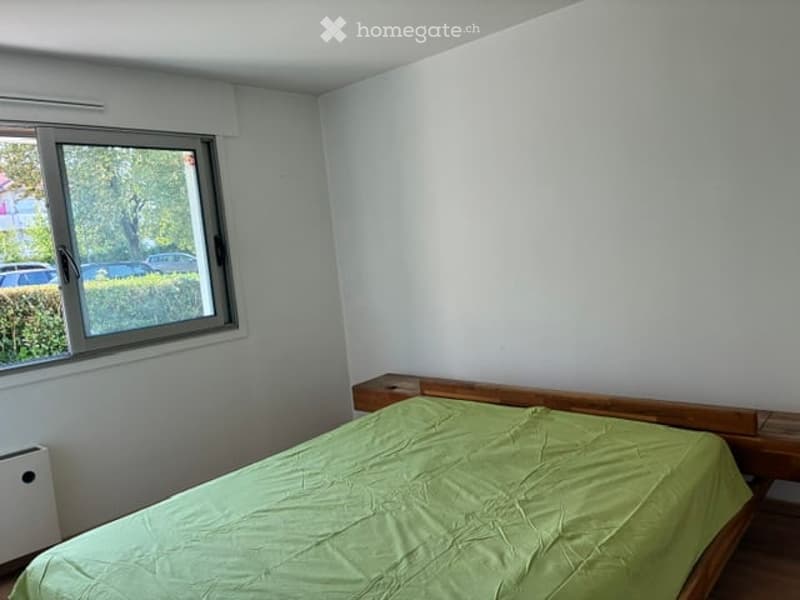 A simple bedroom with a wooden bed and green bedspread, large window with blinds, and white walls.