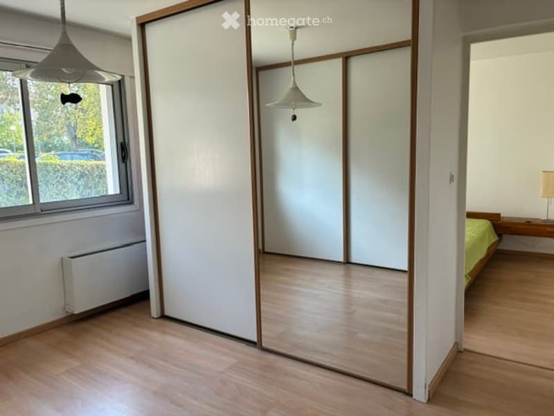 A furnished bedroom with a bed, nightstand, and lamp. The room features wooden flooring, large windows with a view of greenery, and a sliding closet with mirrored doors.