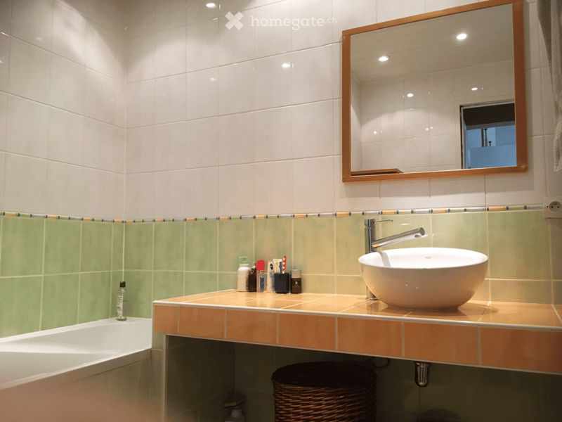 Modern bathroom with white tiles, a large mirror with wooden frame, a sink with a modern faucet, and a bathtub. Toiletries and a wicker basket are on the counter.
