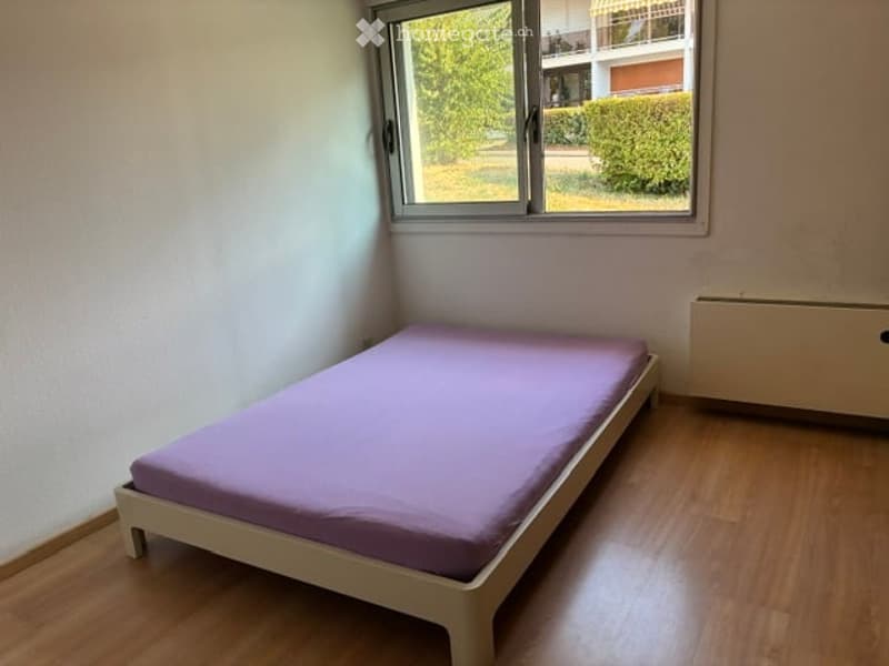 Minimalist bedroom with a single bed, wooden flooring, and a window offering a view of greenery outside.