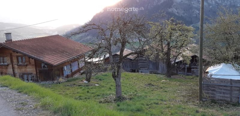 A rustic wooden house with a red-tiled roof and a chimney, surrounded by a grassy yard with trees and a small wooden fence.