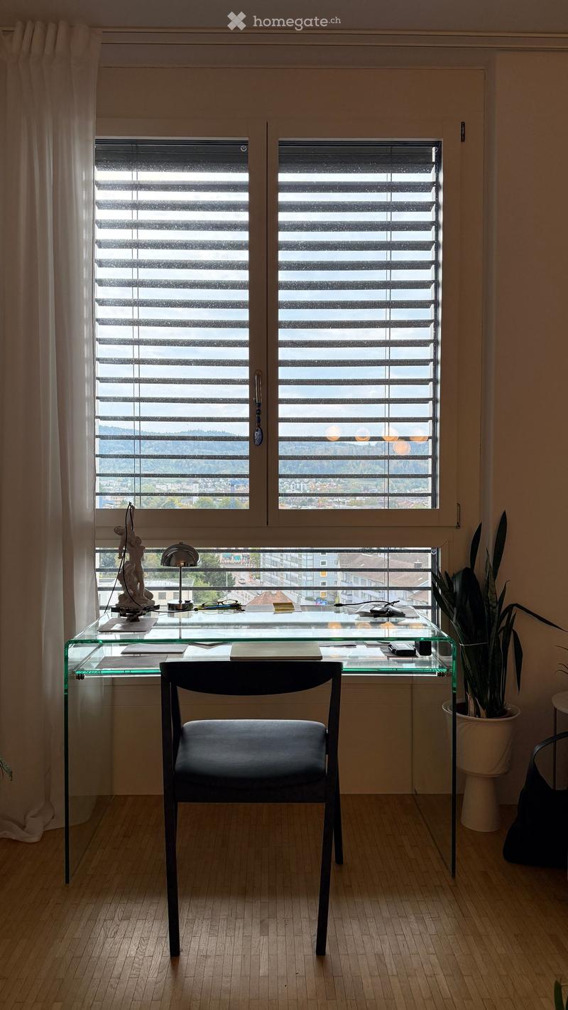 A modern, minimalist room with a glass desk, a black chair, a lamp, a potted plant, and a decorative statue. The room features large windows with blinds and a scenic view of the city.