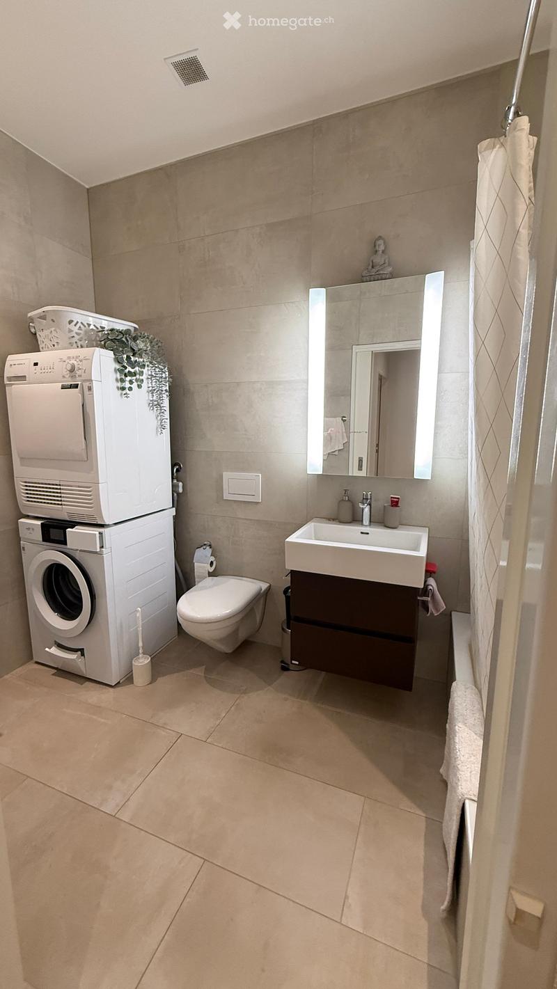 Modern bathroom with beige tiles, a white sink with a mirror, a toilet, a washing machine, and a shower area with a curtain.