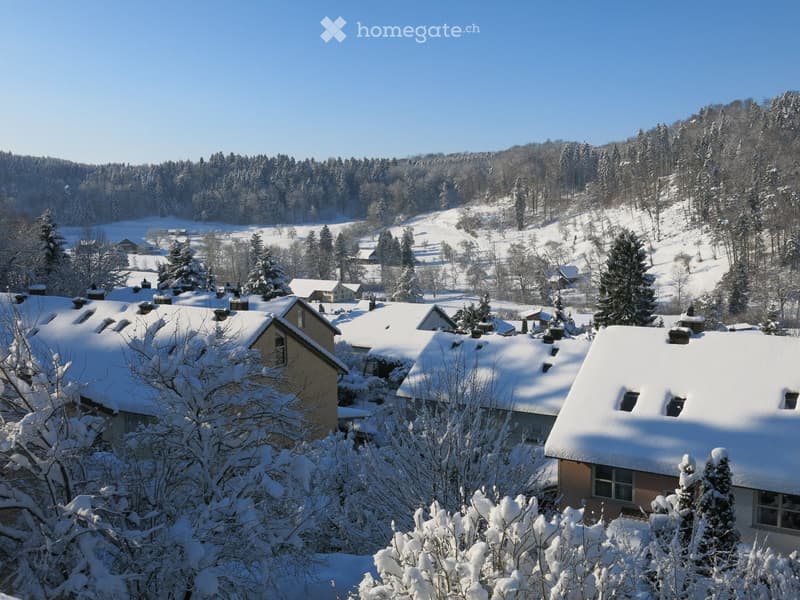 A snow-covered village with multiple houses and trees in a forested area under a clear blue sky.