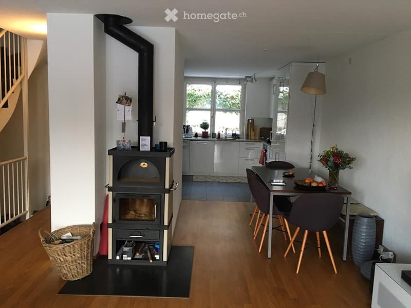 A modern living space with a wood stove, dining table with chairs, and an open kitchen area with cabinets and a sink.