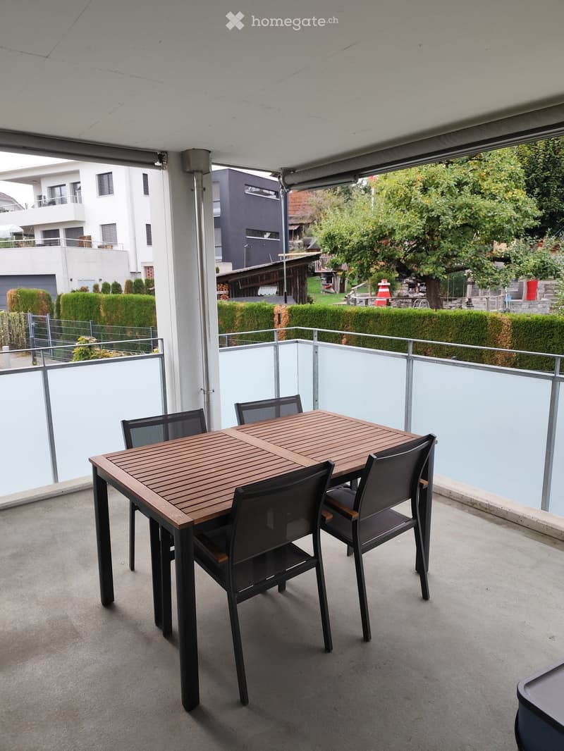 Balcony with a wooden table and four black chairs, surrounded by a glass railing and overlooking a landscaped garden area.