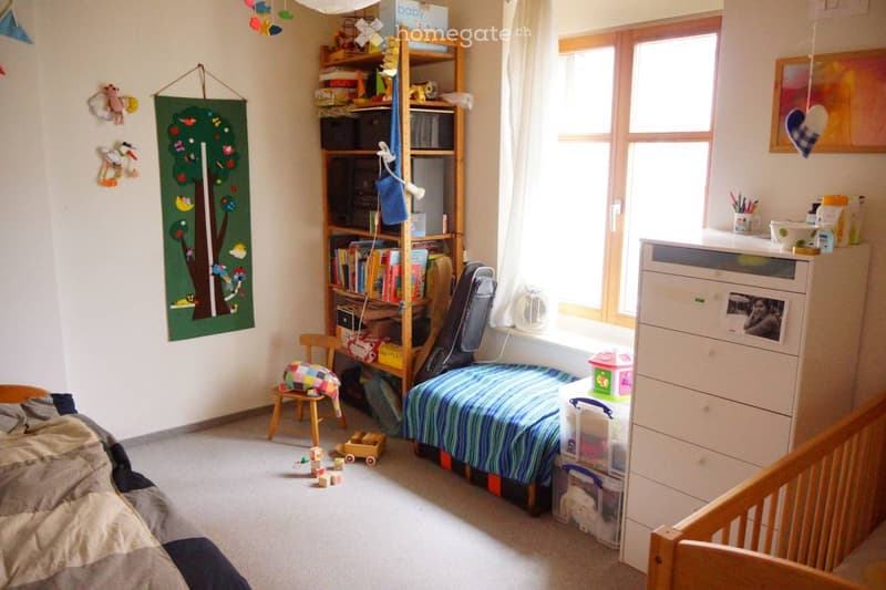 A well-lit children's bedroom with a single bed, a small chair, a bookshelf filled with books and toys, a small table with a cup, and a crib visible in the foreground.