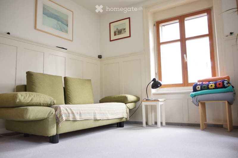 A cozy living room with a green sofa, small white table, black lamp, and a wooden stool with folded blankets.
