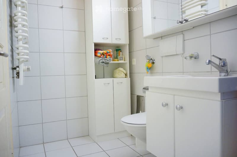 Modern bathroom with white tiles, a sink with a chrome faucet, a toilet, a mirrored cabinet, and a towel rack with towels.