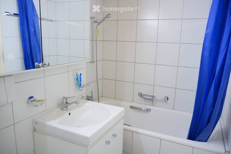Bathroom with white tiled walls, a white sink with a chrome faucet, a bathtub with a blue shower curtain, a mirror, and a toothbrush holder on the wall.