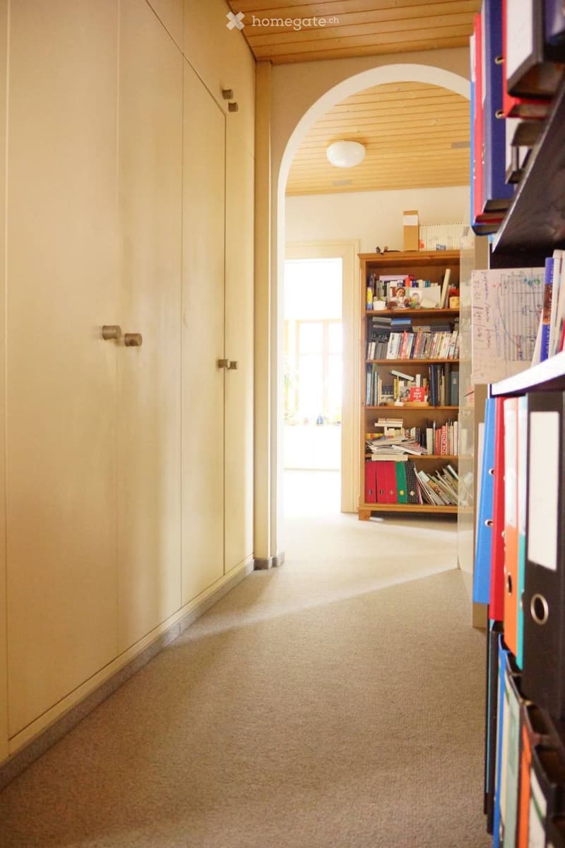 Carpeted hallway with light-colored walls, wooden ceiling, and a wooden bookshelf filled with books and binders on the right side.