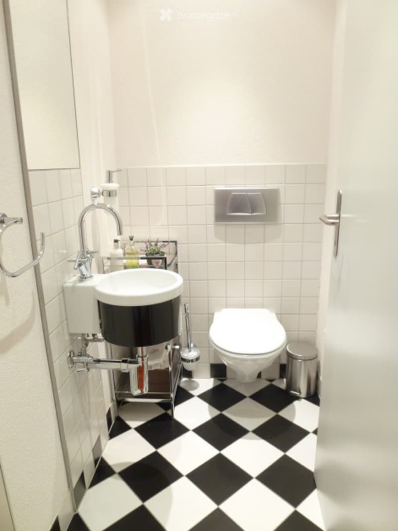Modern bathroom with white tiled walls, black and white checkered floor, white sink with chrome faucet, toilet, and towel ring.