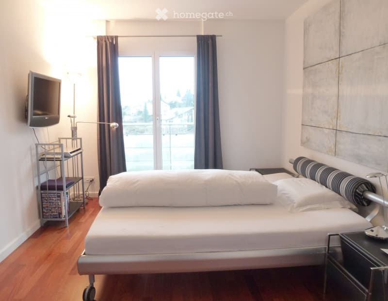 Modern bedroom with a large window and balcony access, featuring a bed, nightstand, lamp, and a TV on a stand.