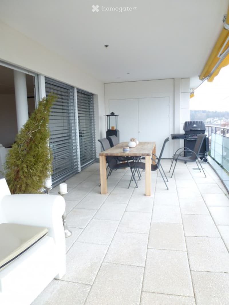 Modern balcony with tiled floor, large windows, dining table with chairs, grill, and a small tree.