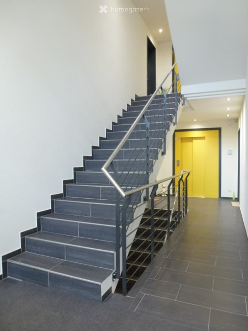 Modern hallway with a staircase leading upwards, featuring grey tiled steps, metal railings, and a bright yellow door at the end.