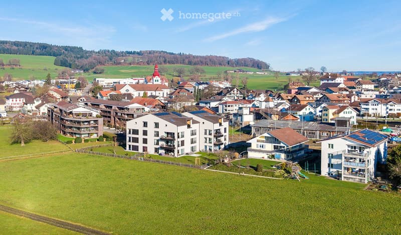 Aerial view of a residential area with multiple buildings, including apartment complexes and houses, surrounded by green fields and a small village with a church in the background.