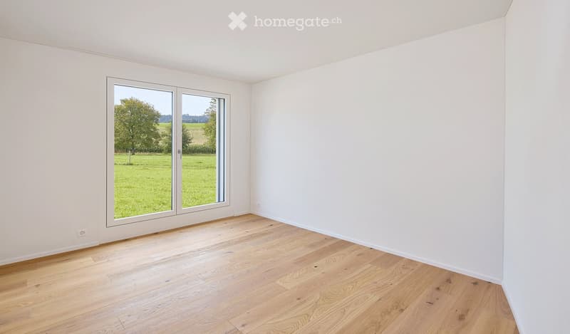 Empty room with light wooden flooring and white walls, featuring a large sliding glass door opening to a green outdoor area.