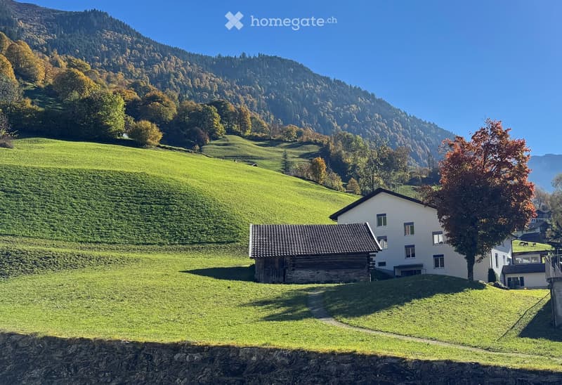A two-story house with a tiled roof and white exterior, situated on a grassy hillside with a wooden outbuilding nearby. The surrounding area features lush green fields and a mountainous backdrop.