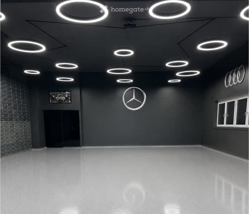 Empty room with dark walls, a polished concrete floor, and circular ceiling lights. Features a large Mercedes-Benz logo on one wall and a framed picture on another.