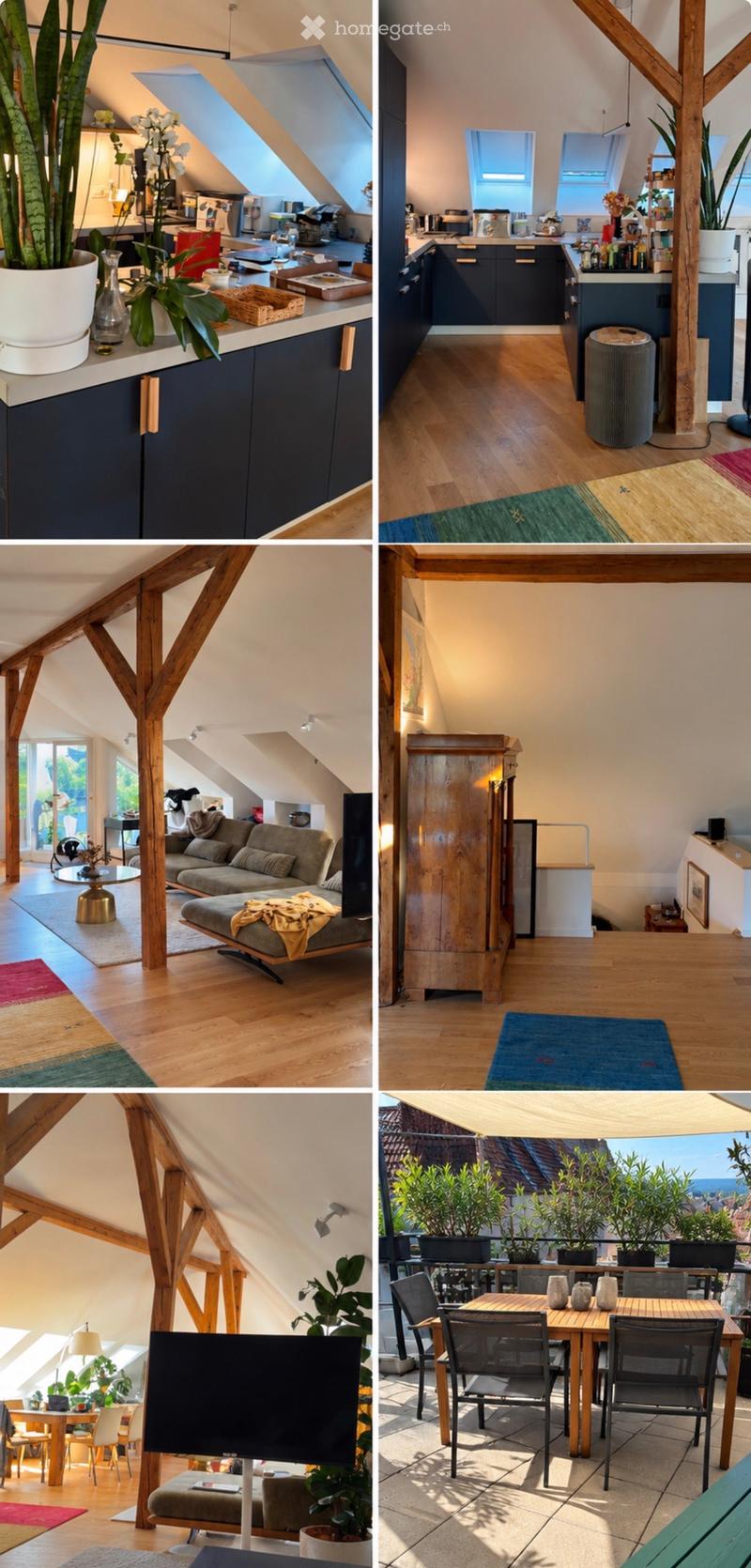 A modern attic apartment with exposed wooden beams, large windows, and skylights. Features include a kitchen with dark cabinets, a living area with a sofa and coffee table, a dining area with a wooden table and chairs, and a balcony with outdoor seating a