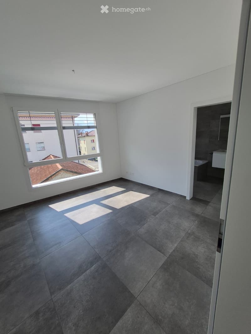 Empty room with grey tiled floor, white walls, and large windows showing a view of neighboring buildings. Sunlight streams in, casting shadows on the floor.
