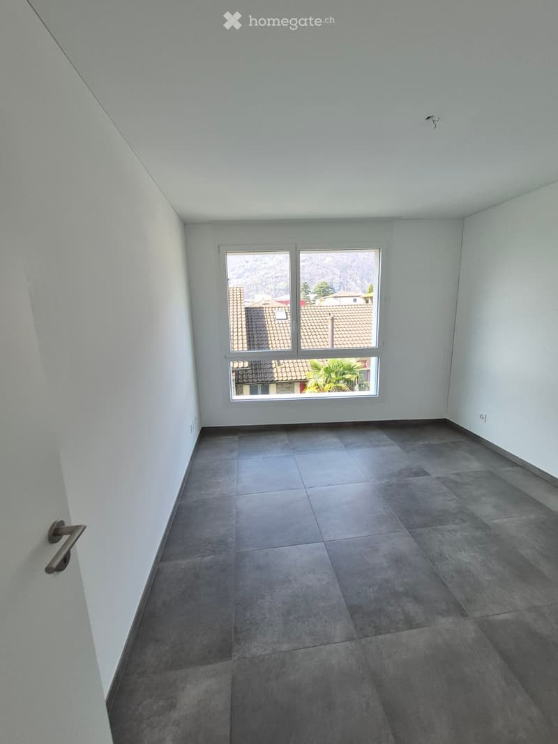 Empty room with grey tiled floor, white walls, and a large window offering a view of rooftops and mountains.