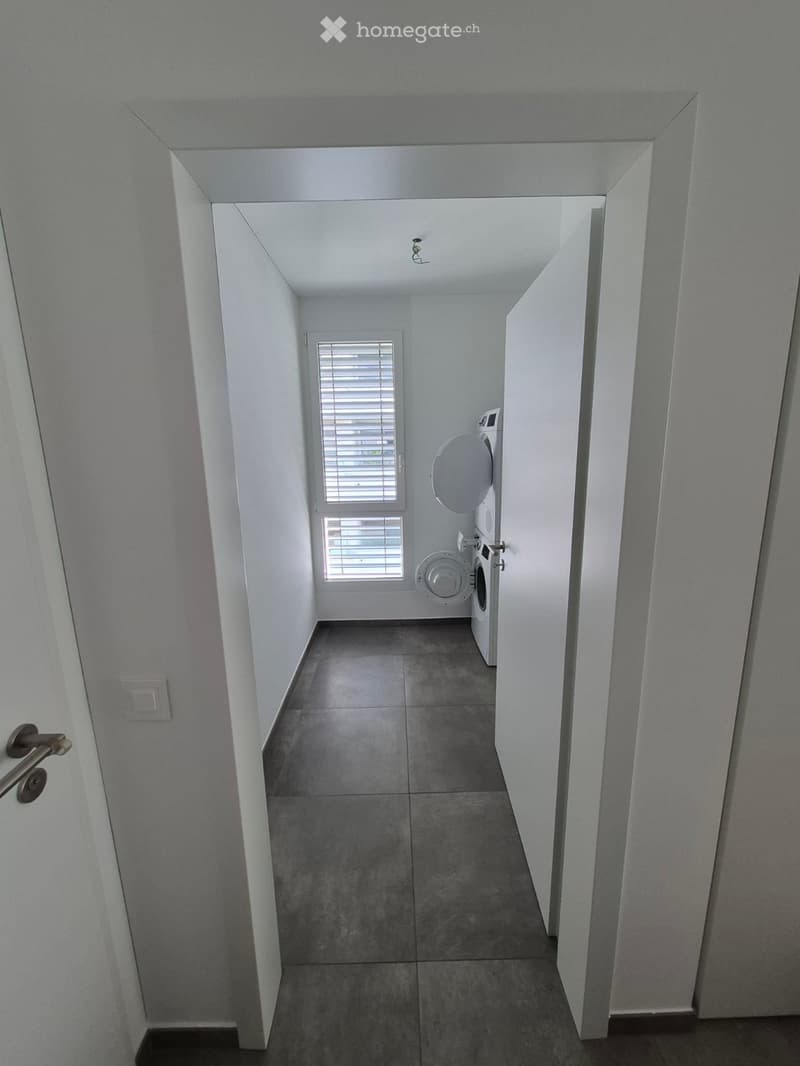 A narrow, well-lit hallway with tiled flooring and white walls, leading to a room with a washing machine and dryer visible at the end.