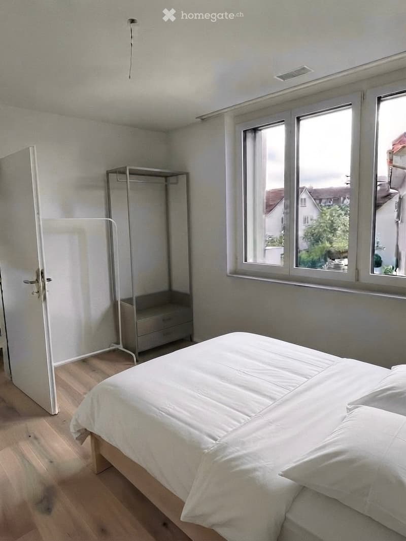 A modern, minimalistic bedroom with a white bed, a wardrobe, and a clothing rack. The room features wooden flooring and large windows offering a view of the neighborhood.