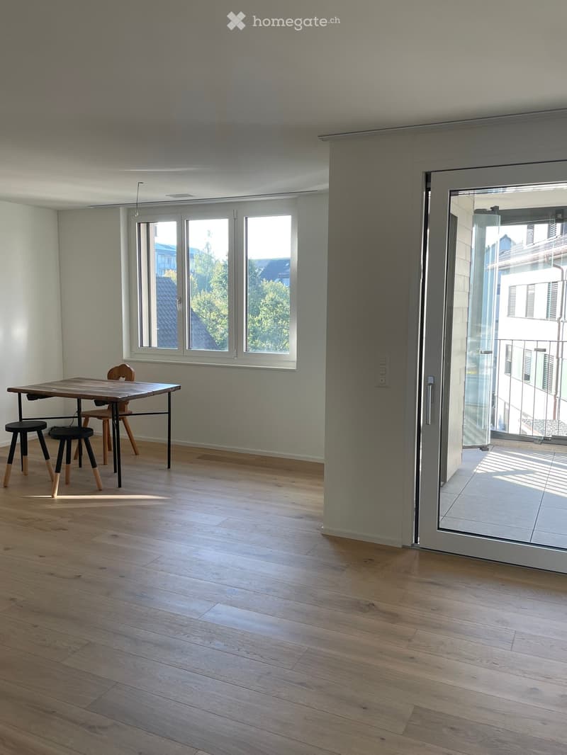 Bright, minimalist room with wooden flooring, large windows, and a sliding glass door leading to a balcony. Contains a small wooden table and two chairs.