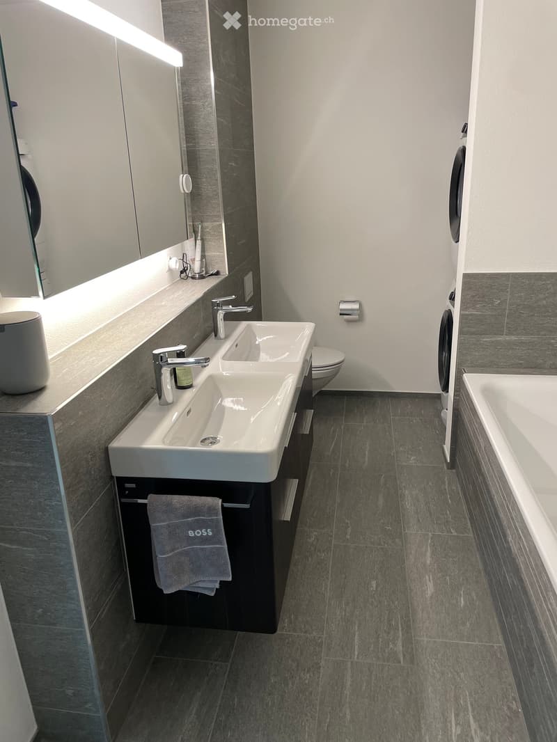Modern bathroom with double sink, mirror, bathtub, toilet, and towel on cabinet.