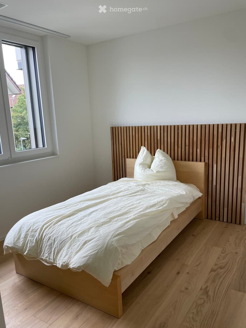 A modern bedroom with a wooden bed, white bedding, and a sleek headboard. The room features light wooden flooring and a large window with a view of greenery.