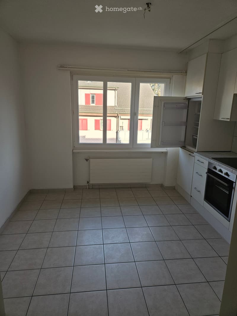 Empty kitchen with tiled floor, white cabinets, and a window overlooking a neighboring building. No appliances or furniture are present.