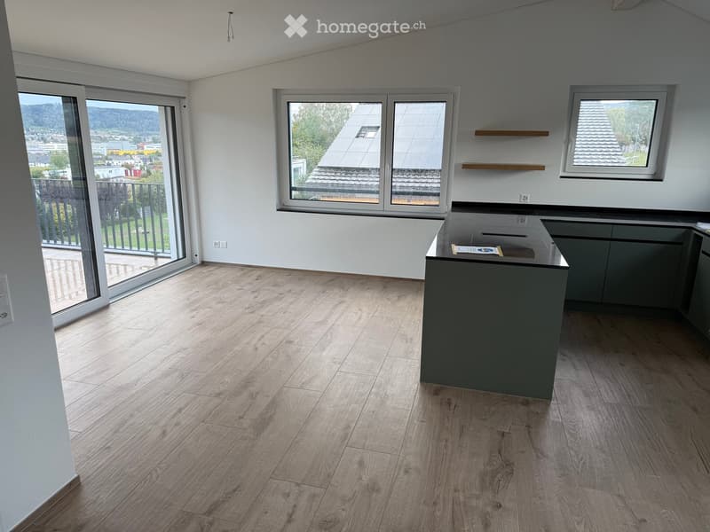 Empty kitchen with wooden flooring, grey cabinets, and a countertop. Large windows and a sliding glass door lead to a balcony.