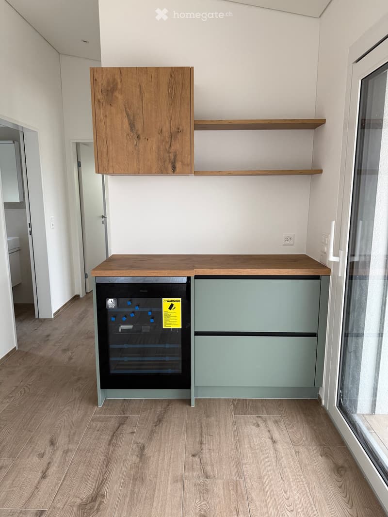 Modern kitchen with light wood cabinets, a countertop, and a dishwasher. The space features wooden flooring and a large window on the right side.