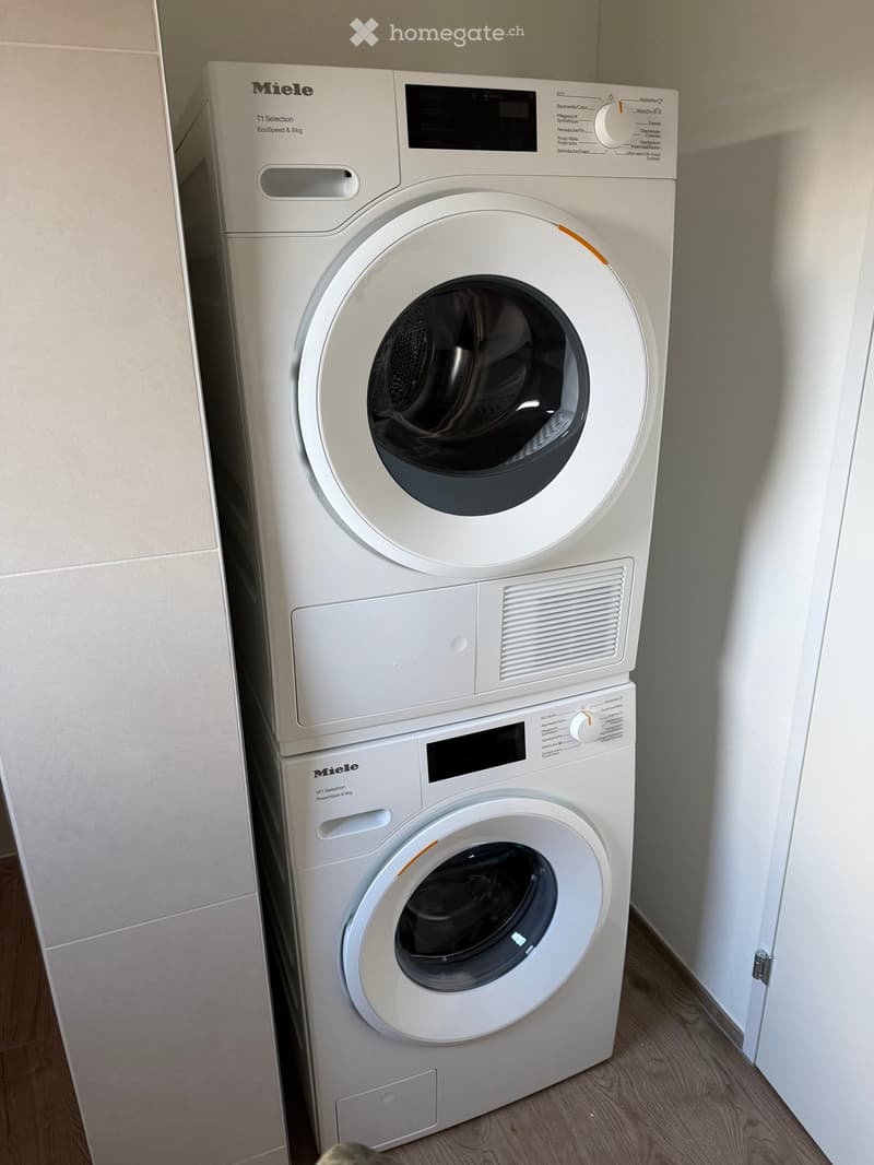 White Miele washing machine and dryer stacked vertically in a bathroom with light-colored walls and wooden flooring.