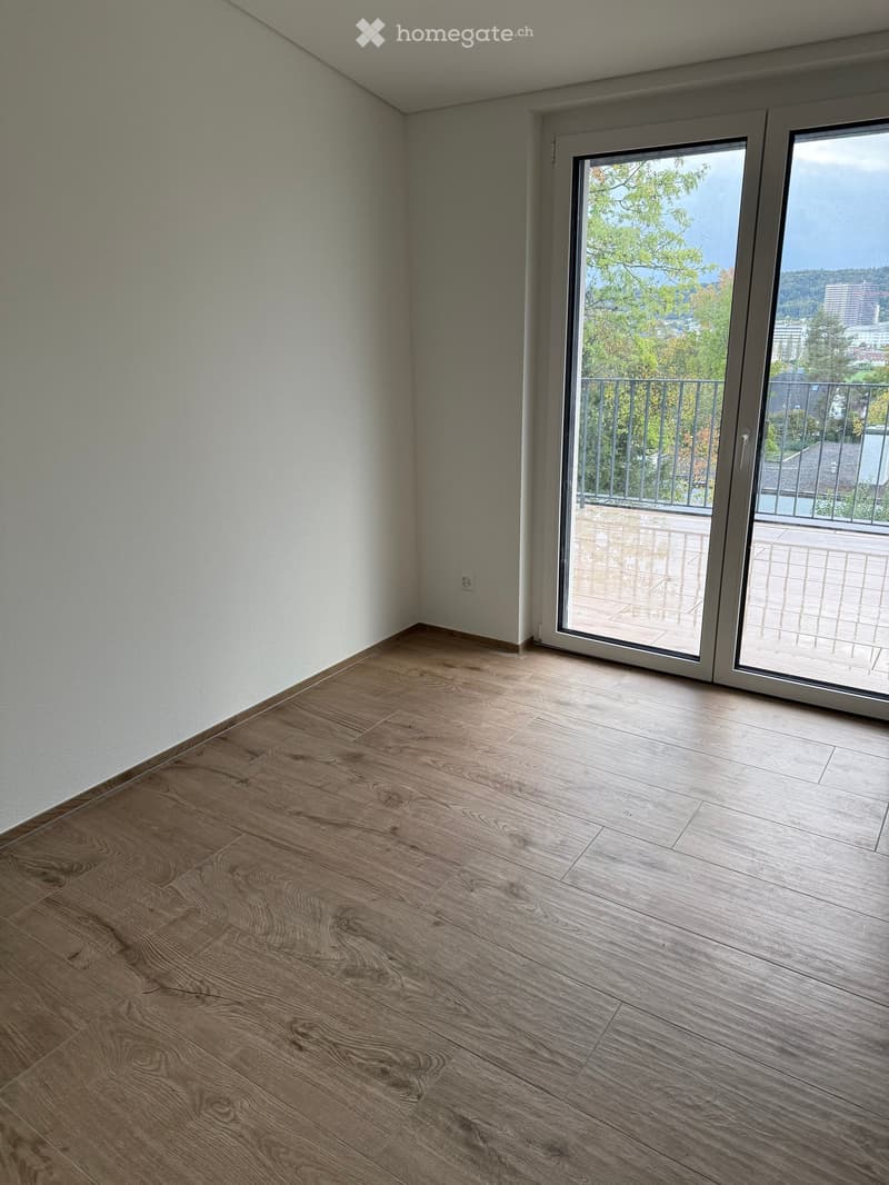 Empty room with wooden flooring and large sliding glass doors leading to a balcony with a view of trees and cityscape.
